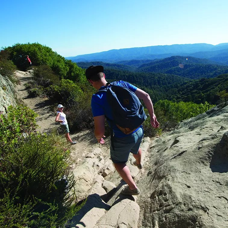 People hiking on a trail that drops down over the redwood covered valley of Castle Rock State Park